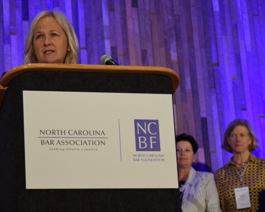 Patti Ramseur is a white woman with shoulder-length blond hair. She is wearing small pearl earrings. She stands at a podium, and from the angle, only her head is visible. In front of the podium is a white sign with the NCBA logo in blue. In the lower right of the photo, a few feet away from the stage, stand Shelby Benton and Caryn McNeill, two past presidents. Shelby Benton is a white woman with short dark hair, who is a past president. Shelby is wearing a dark blouse and a white and pale blue pinstripe jacket with a multi-layered strand of pearls. Caryn McNeill, stands to the right of Shelby, and Caryn has short light brown hair and is wearing it in a bob, and she is wearing a gold and black button down dress with gold jewelry. The backdrop behind the stage is a periwinkle blue that appears like long lit streamers. 