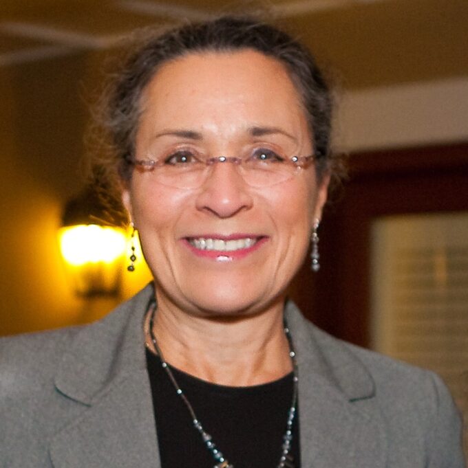 Arlinda Locklear is a Lumbee woman with dark brown hair and brown eyes, and she is wearing clear glasses with a gold trim on the nosepiece and sides of the glasses. She is wearing a black blouse with a pale grey jacket, and a pale grey beaded earrings and a matching necklace. She is pictured smiling with a softly lit beige room in the background.