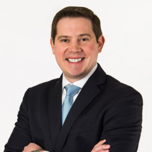 Andrew Atkins is a white man with brown hair. He is pictured in a white shirt, pale blue tie, and black jacket. He is pictured smiling and folding his arms, and he stands against a white background.
