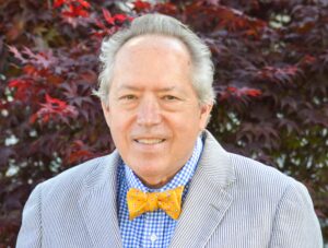 Brad Schultz is a white man with grey hair and dark eyes. He is wearing a blue and white checkered shirt yellow bowtie with red, blue and white squares and a grey and white pinstripe jacket. He is photographed standing with red foliage in the background. 