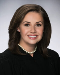 Judge Carrie Vickery is a white woman with brown eyes and shoulder-length dark brown hair, and she is pictured wearing a black judge’s robe with a white pearl necklace and pearl stud earrings. She is photographed against a white background.