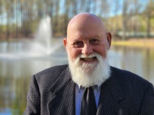 Cecil Whitley is a white man with a white beard, and he is wearing a blue shirt, black tie and black jacket with small white specks. He is pictured smiling and standing outside in the sun with a blurred fountain behind him. 