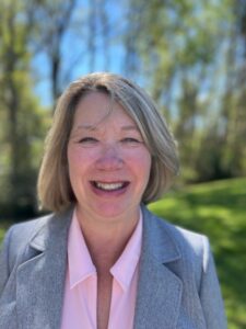 Debbie Sandlin is a white woman with short, light-blonde hair. She is wearing a pink button-down shirt and a grey jacket, and she is pictured smiling outdoors with a blurred green field and trees behind her. 