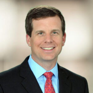 Ben Edwards is a white man with dark brown hair and dark eyes. He is wearing a light blue shirt, red tie and black jacket. He is smiling, and the background behind him is blurred.