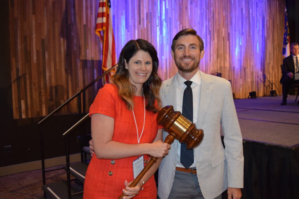 Lisa Williford stands next to Will Quick. Lisa holds a large brown gavel in her right hand. Lisa is a white woman with medium length brown hair with lowlights. She is wearing a bright orange short-sleeve dress with brown buttons. Will Quick is a white man with light brown hair and a beard and mustache. He is wearing a white shirt, navy tie, and white and grey striped suit jacket, grey pants and a brown belt. Lisa and Will stand with a stage behind them, which has a shiny silver backdrop, and the American flag appearing just behind Lisa's right-hand shoulder. 