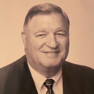 Gerald Thornton is a white man with dark hair. He is pictured in a black and white style photo with a sienna tint, and he is wearing a white shirt, dark jacket, and black and white tie.
