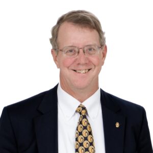Robert H. (Bob) Haggard is a white man with light brown hair. He is wearing a white shirt with a yellow and black patterned tie, a black jacket, and a gold pin on the right-side lapel. He is wearing wire-rimmed glasses and is pictured smiling, and he stands against a white background.
