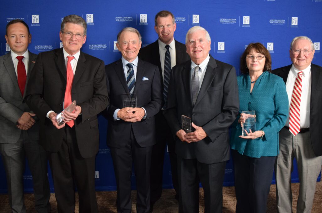 Front from left, Hall of Fame inductees are pictured: Richard Boyette, Kenneth Greene, Jody Moore and Sharon Parker. Back: Executive Director Jason Hensley, President Jon Heyl and Ed Gaskins, who presented the awards on behalf of the Senior Lawyers Division. Inductee Gerald Thornton, who was unable to attend, is pictured below. Richard Boyette is a white man with grey hair and wire-rimmed glasses, and he is wearing a white shirt, coral tie, and a black jacket; he is pictured smiling. Kenneth Greene is a white man with grey and white hair, and he is wearing a pale blue shirt, a navy and grey striped tie, and a black jacket; he is smiling. Jody Moore is a white man with white hair, and he is wearing a cream-colored shirt, a grey tie, and a black jacket; he is pictured smiling. Sharon Parker is a white woman with dark reddish-brown hair, and she is wearing brown glasses with a teal button-down blouse, and black pants. Pictured in the back row are Jason Hensley, a white man with brown hair who is wearing a red tie and a grey suit with an Americn flag pin on his lapel, Jon Heyl, a white man with brown hair who is wearing a navy blue and grey striped tie and black suit, and Ed Gaskins, a white man with grey hair and wire glasses who is wearing a white shirt, red and white striped tie, black jacket and grey pants. 