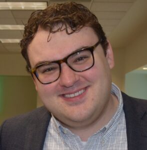 Jared Smith is a white man with brown curly hair and brown glasses. He is pictured with his head tilted to the left and smiling, and he is wearing a grey button down shirt and dark grey jacket.