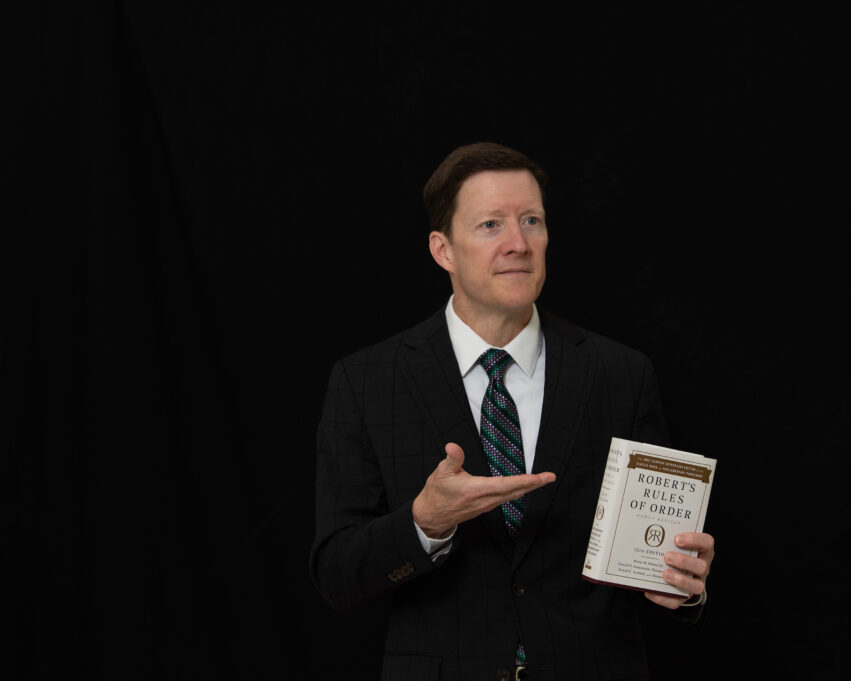 In this image, Jim Slaughter, a white man with dark brown hair, stands against a black background with his hand extended towards the book "Robert's Rules of Order." The book is cream-colored with a brown border on the top. He is wearing a white shirt, dark suit, and tie with dark teal and silver diagonal stripes.