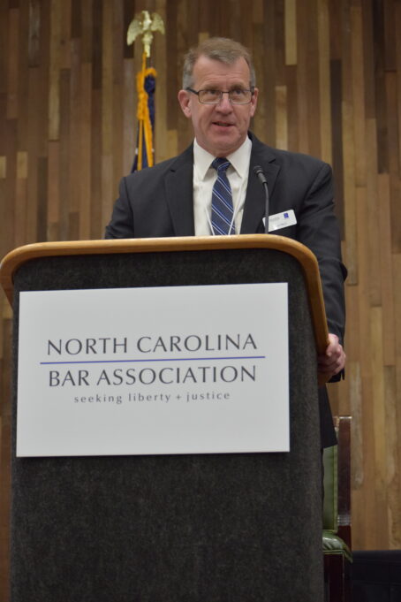 President Jon Heyl introduces Judicial Independence Symposium participants. Heyl is a white man with brown hair who is wearing a black suit and a navy-blue and grey striped tie. He stands in front of a podium that has a white sign that reads “North Carolina Bar Association” in black text and all caps, and “seeking liberty + justice in lowercase beneath. Heyl is standing in front of a gold background and the American flag is visible behind him.