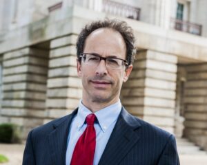 Jonathan Blumberg is a white man with brown hair. He i wearing black glasses, a white shirt, a red tie and a black jacket. He is photographed with a beige building behind him.