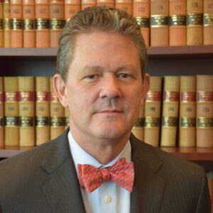 Judge Graham Shirley is a white man with brown hair and dark eyes. he is wearing a coral bowtie, a white shirt and a brown jacket. He stands in front of a bookcase with gold legal books.