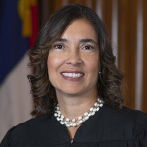 Justice Anita Earls is woman with brown, shoulder-length hair and brown eyes. She is pictured wearing a black robe and a double strand of pearls, and she is smiling. She stands in front of a wood-paneled wall with the state flag in the background.