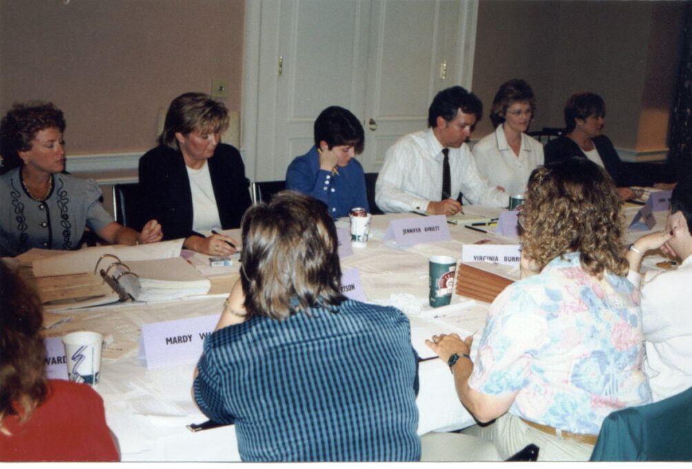 This image captures a meeting of the Legal Assistants Division being held in Winston-Salem.Ten indviduals sit at a long table in a hotel meeting room. Six of the individuals appear sitting next to each other on one side of the table, near the top of the photo, so that their faces are visible. The first woman, who is white, is wearing a grey button-down jacket, and she has curly brown hair. The second woman, who is white, has brown hair, and is wearing a black jacket and white shirt. The third woman, who is white, has short dark hair, and she is wearing a blue button-down shirt. The fourth person is a man, who is wearing a white button-down shirt and dark tie. The fifth person is Grace, who has light brown hair, wire-rimmed glasses, and is wearing a white button down shirt. The sixth person is a woman with short brown hair and a white shirt and black jacket. A binder is visible on the table, as are name tags of the people sitting there. There are four people on the opposite side of the table, pictured with their backs to the viewer. One woman has brown hair with a rust-colored shirt, another woman has short brown hair and is wearing a blue plaid shirt, another woman with curly hair has a pale floral shirt with blue and pink flowers, and the fourth woman has short, dark hair and is wearing a white sweater. 