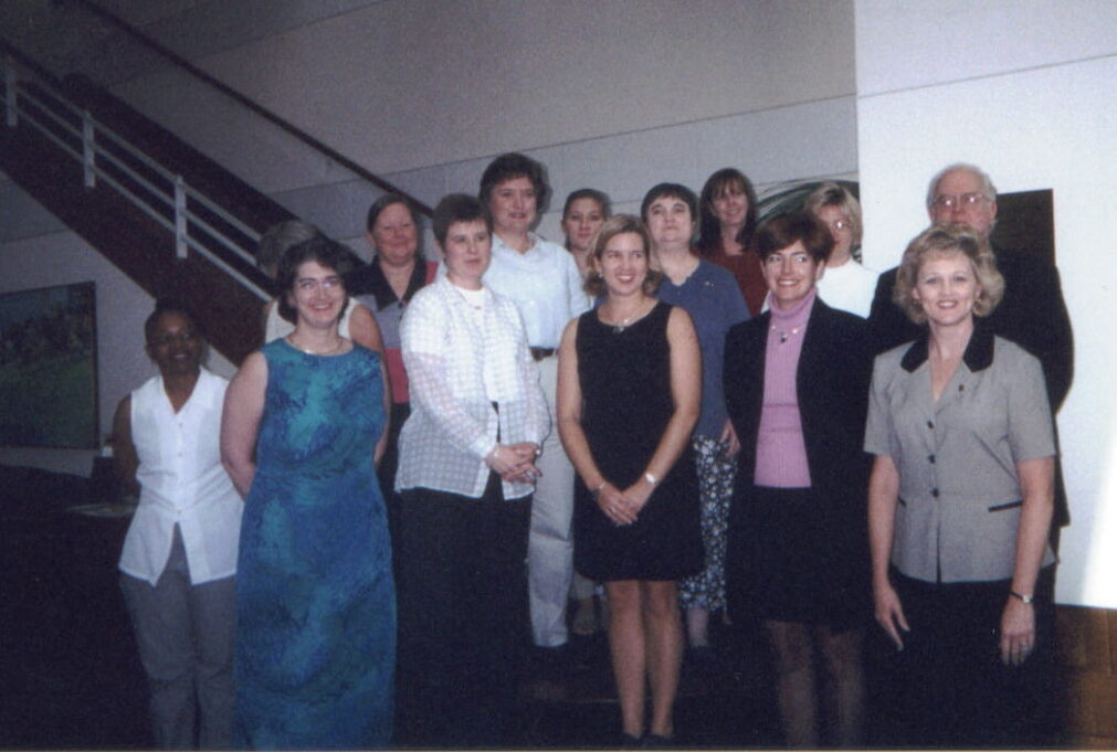 In this photo, fourteen people stand in front of the stairway at the bar center in Cary. Left to right, the individuals are pictured and you see a Black woman, who stands on the left, and she has dark hair and is wearing a white button-down shirt, a woman with short brown hair and glasses who is wearing a blue dress, a woman behind her with grey hair and a white shirt whose face is obscured, a woman with brown hair ina ponytail and a grey and orange colorblocked dress, a woman in the front with short brown hair and a white button-down shirt, a woman in the back with dark, short brown hair and a white-button down shirt, a woman behind her with brown hair in a ponytail, a woman in front with short light-brown hair and a black dress, a woman with short dark hair in a blue shirt and patterned pants, a woman with brown hair in the back in a rust-colored shirt, a woman with short brown hair in the front and who wears a purple turtleneck with a black jacket, and Grace, who stands on the far right and is wearing a light grey short-sleeved suit jacket with a black color. Grace's hair is short, wavy and blond. A man stands behind her, and he has grey hair, glasses, and a black jacket on. 