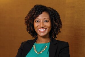 Jocelyn Mallette is a Black woman with brown eyes and curly, shoulder-length hair. She is wearing a teal blouse, gold necklace, and black suit jacket, and she is standing against a warm camel-colored wall. 