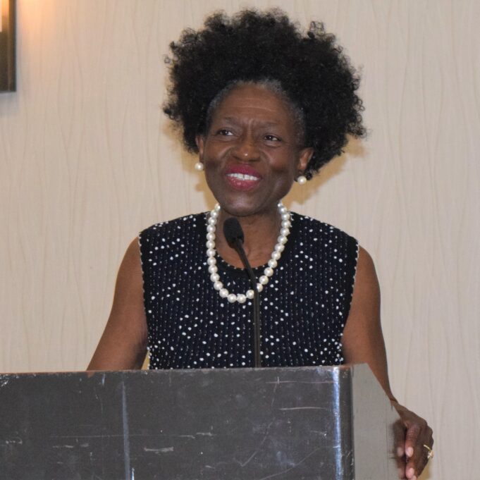 Margaret Dudley stands at a grey podium and in front of a white background. Margaret is a Black woman with black hair, and she is pictured smiling and wearing a black dress with a pearls woven into the fabric, as well as a pearl necklace, pearl earrings and a pearl bracelet. 