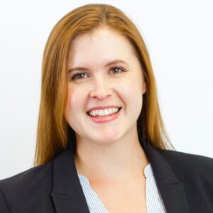 Martha Bradley is a white woman with brown eyes and red hair. She is wearing a white shirt and black suit jacket. She stands against a white background.