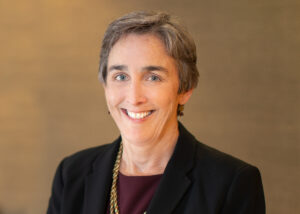 Mary Nash Rusher is a white woman with grey eyes and short brown hair with grey highlights. She is pictured wearing a maroon blouse and black suit jacket, and she is smiling.