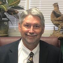 Bob Laney is a white man with grey hair and a grey mustache and beard, and he is smiling. he sits in a maroon office chair with a windowsill and blinds behind him. On the windowsill sits a plant on his left and a decorative quail to the fair right. Bob is wearing a white shirt, black suit, and black and gold tie.