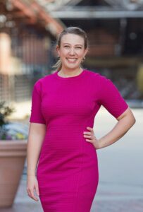 Michele Livingstone is a white woman with blond hair. She is pictured with her hair in a ponytail and wearing a bright pink dress, with her left hand on her hip, and she stands in a courtyard.