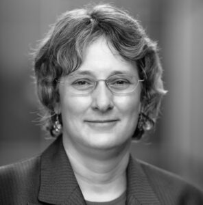 Nan Hannah is a white woman with wavy brown hair. She is pictured wearing wire-rimmed glasses and smiling with her mouth closed. She is pictured in a greyscale photo and is wearing a dark blouse with a dark suit jacket.