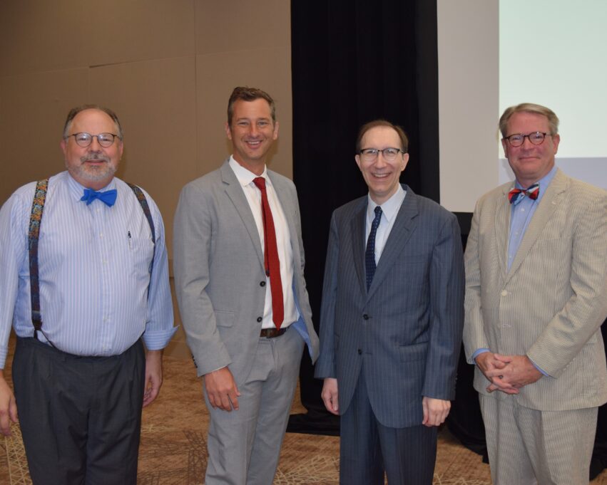 From left, Joe Stewart, Christopher A. Cooper, Mitch Kokai and J. Michael Bitzer. Joe Stewart is a white man with grey hair and a grey beard. He is smiling and wearing black round glasses, a blue and white striped button-down shirt, a bright blue bowtie, dark green and gold patterned suspenders, and grey pants. Christopher A. Cooper is a white man with brown hair, and he is smiling and wearing a white button-down shirt, red tie, and pale grey suit. Mitch Kokai is a white man with dark brown hair, and he is also smiling. He is wearing black glasses, a white button-down shirt, a dark tie, and a grey and blue pinstriped suit. Michael Bitzer is a white man with light blond hair, and he is also smiling. Michael wears a brown circular glasses, a blue button-down shirt, off-white pinstriped suit, and a bowtie that is red, cream, and pale blue. They stand near the front of the stage, and the projector is visible behind Michael.