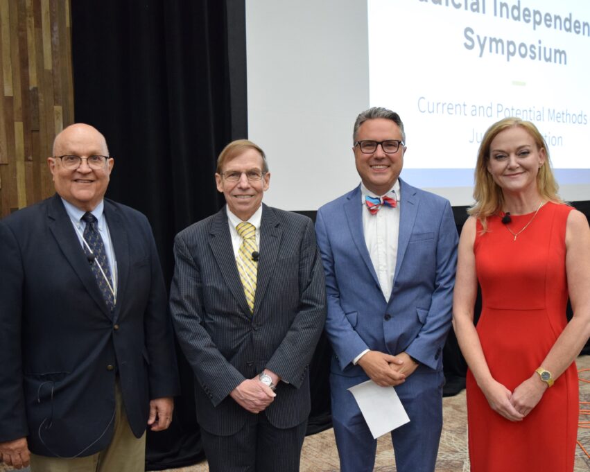 From left, James C. Drennan, Robert H. (Bob) Edmunds Jr., Tim R. Boyum and Jeanette Doran. James C. Drennan is a white man with black glasses, and he is wearing a blue button-down shirt, dark blue tie, dark blue suit jacket, and khaki pants, and he is smiling. Robert H. (Bob) Edmunds Jr. is a white man with light brown hair and black glasses, and he is smiling and wearing a white button-down shirt, yellow tie with blue stripes, and a black and grey pinstriped suit. Tim R. Boyum is a white man with black glasses, and he is wearing a white button-down shirt, medium blue suit, and a red, blue and white bowtie. Jeanette Doran is a white woman with blond, shoulder-length hair, and she is smiling with her mouth closed. Jeanette is wearing an orange-red dress and gold necklace. The participants stand in front of the stage, and the screen is visible behind Robert, Tim and Jeanette.