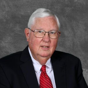 Richard Doughton is a white man with white hair and blue eyes. He is wearing black wire-rimmed glasses, a white shirt, a black jacket, and a red and white diagonally striped tie.
