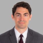 Stephen Bell is a white man with curly black hair and blue eyes. He is wearing a white shirt with a black tie and a grey jacket. He stands in front of a grey background and is smiling with his mouth closed.