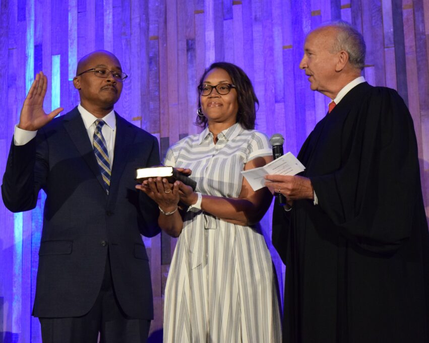 Clayton Morgan stands with his wife Kimberly and Chief Justice Paul Newby, who administers the oath of office. Clayton Morgan has his right hand raised. Clayton is a Black man with brown hair, and he is wearing glasses, a dark blue suit, a white shirt, and a grey and white plaid tie. Kimberly holds the Bible. Kimberly is a Black woman with shoulder-length black hair and dark glasses, and she is wearing a grey and white striped button-down dress. Chief Justice Newby is wearing a white shirt with a red tie and a dark suit. The three individuals stand in front of a background with gold and silver streamers.