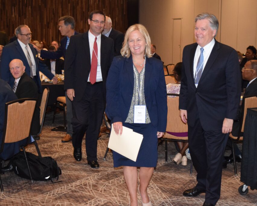 Immediate Past President Mark Holt ushers President-elect Patti Ramseur to the stage, followed by the additional past presidents in attendance at the NCBA Annual Meeting. Patti Ramseur stands next to Mark Holt. Patti is a white woman with shoulder-length blond hair, and she is wearing a white blouse with black flowers and detailing, a navy blue suit jacket and matching skirt, cream-colored shoes, and pearl earrings and a silver necklace. Patti is pictured smiling. Mark Holt is a white man with grey hair, and he is wearing a white shirt with a blue tie and a black suit with black shoes, and he is also smiling. Patti and Mark are standing in a large hotel conference room, where other individuals are seated around tables. One past president is also standing behind Patti, and he has dark hair, black glasses, a white shirt, a red tie and a black suit jacket. 