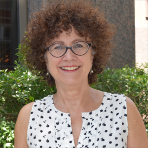 Suzanne Chester is a white woman with curly brown hair. She is pictured wearing dark brown glasses, silver earrings, and a sleeveless white shirt with small black flowers. She stands in front of green foliage and a beige building.