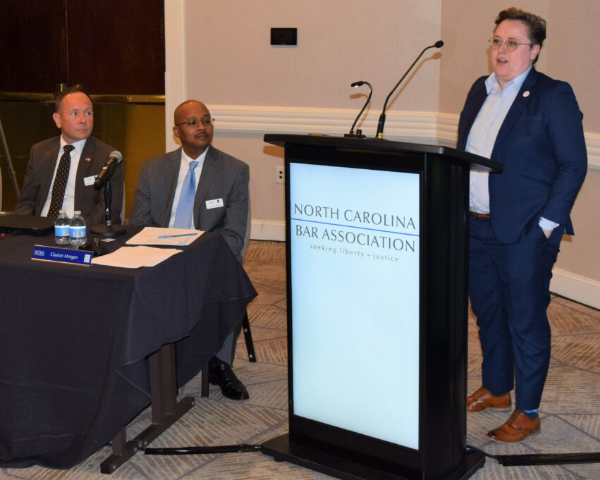 Sarah stands at the podium and Director Jason Hensley and President Clayton Morgan sit at a table on the left.