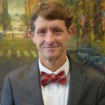 Brian, a white man with brown hair, wears a white shirt, red bowtie, and grey suit.