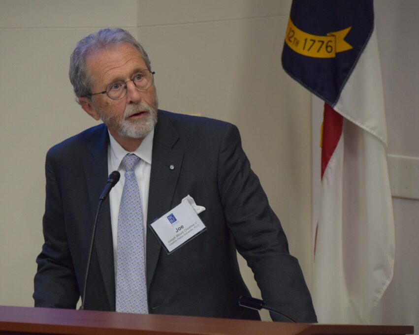 Joe, a white man with grey hair, a beard, and round glasses, wears a white shirt, pale blue tie, and grey suit.