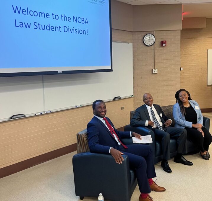 YLD Chair Elect Jonathan Bogues sits with President Clayton Morgan and Sheila Spence at the front of a large auditorium.