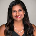 Manisha, an Asian American woman with dark brown hair, wears a black blouse and a white blouse and a pearl necklace.