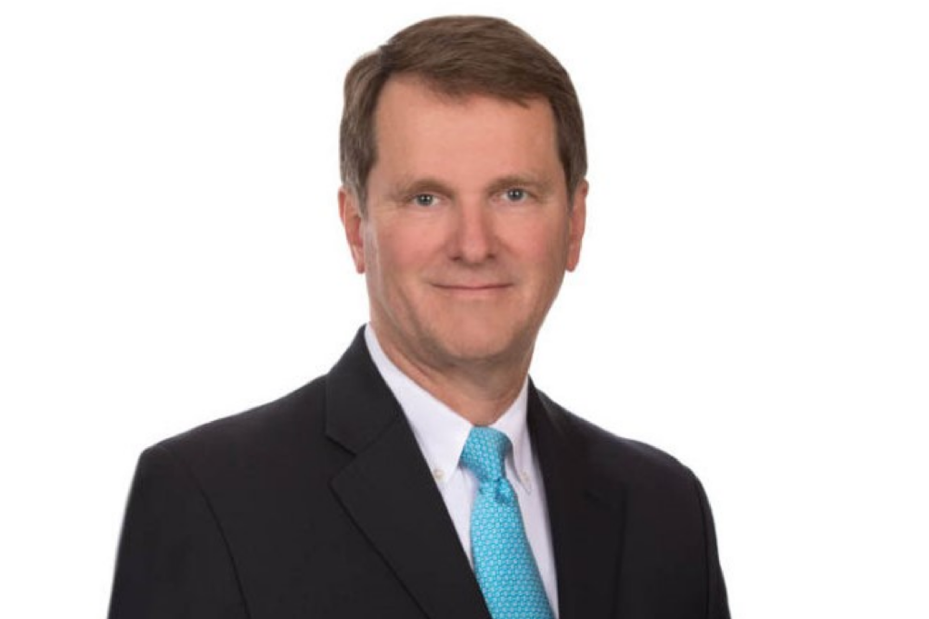 Rudy Ogburn, a white man with brown hair, is pictured wearing a white shirt, blue tie, and black jacket.