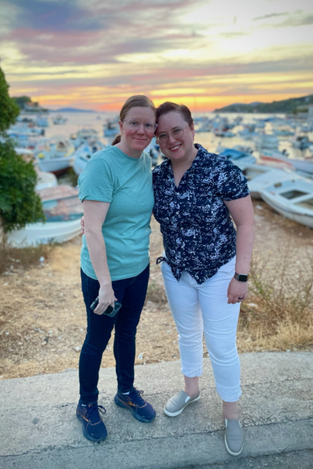 Sarah, a white woman with brown hair, is pictured with wife Kim, a white woman with blond hair. They stand with boats and water in the background.