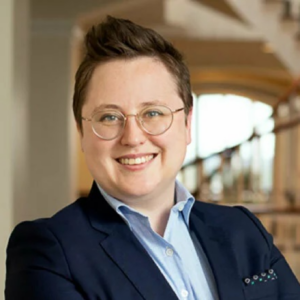 Sarah, a white woman with short brown hair and round glasses, wears a pale blue shirt and navy suit.