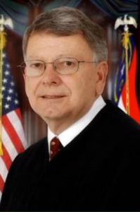 Hunter, a white man with grey hair and glasses, wears a judge's robe, white shirt and red tie with the American flag and NC flag in the background.
