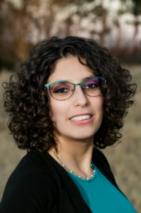 Gadalla, a woman with curly brown hair and teal glasses, wears a teal shirt and black sweater.