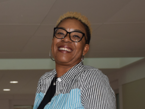 Ebony, a Black woman with short black hair with blond highlights, wears a black blouse and a black, white and teal button-down shirt. She is smiling.