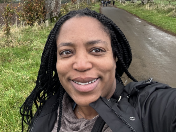 McGhee, a Black woman with black hair, wears a pale grey sweater and a black jacket. She is smiling and standing with a long road, green foliage, and large trees behind her.