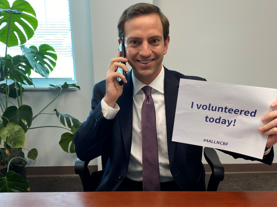 Wilson, wearing a white shirt, purple tie and navy suit, holds a phone to his ear and a sign that says "I volunteered today!"