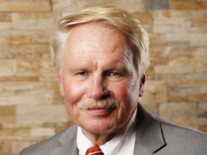 Leonard, a white man with pale blond hair and a mustache, wears a white shirt, red tie, and grey blazer.