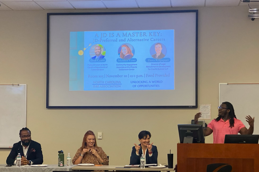 A Black panelist wearing a coral shirt is shown at a podium. Three presenters are seated at a table nearby.