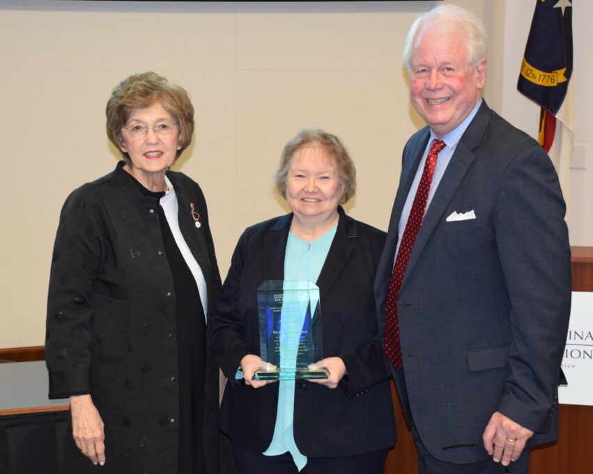 Ann Wall, a woman with blond hair, wears a teal shirt and black jacket and is holding her award. She stands with Elaine Marshall and Bain Jones.